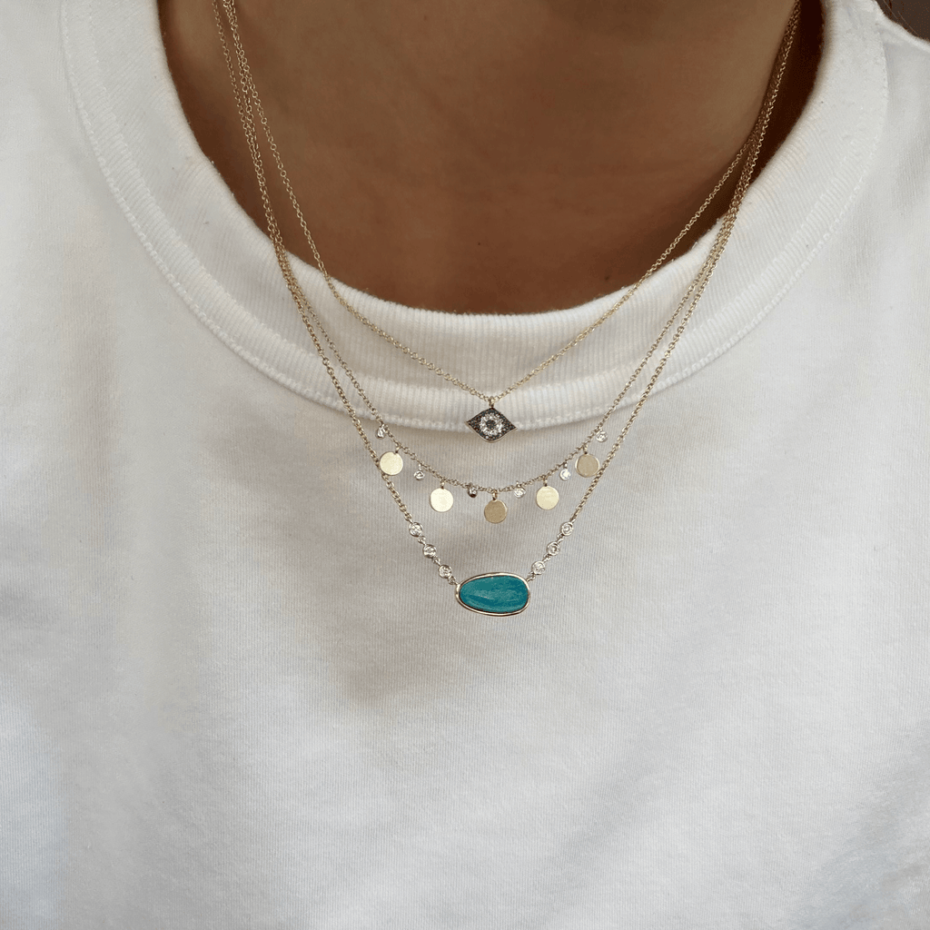 A close-up of layered Meira T 14kt gold necklaces. The necklaces feature gold chain with an evil eye pendant, a second chain with gold disc charms, and a third chain with an oval opal pendant.
