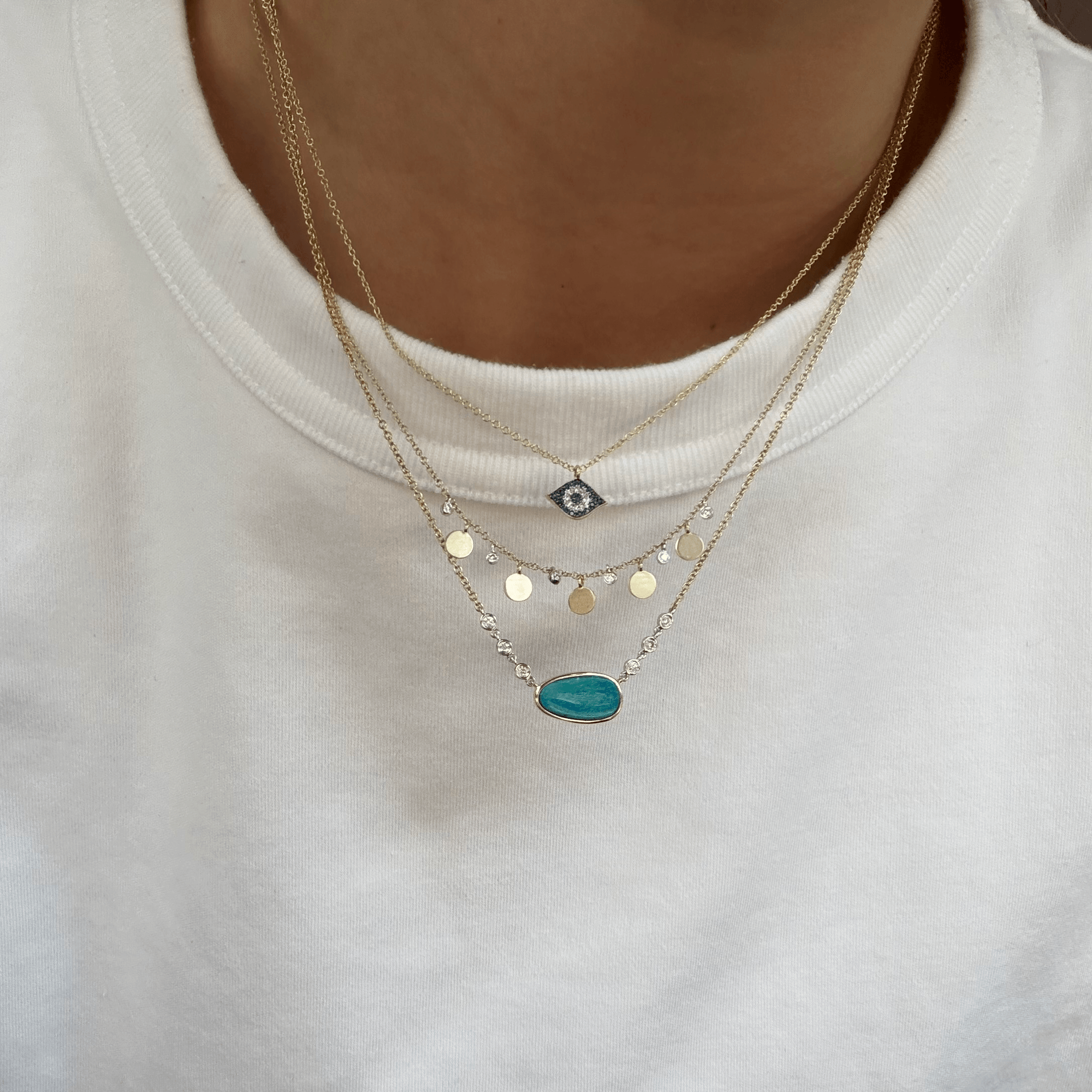 A close-up of layered Meira T 14kt gold necklaces. The necklaces feature gold chain with an evil eye pendant, a second chain with gold disc charms, and a third chain with an oval opal pendant.