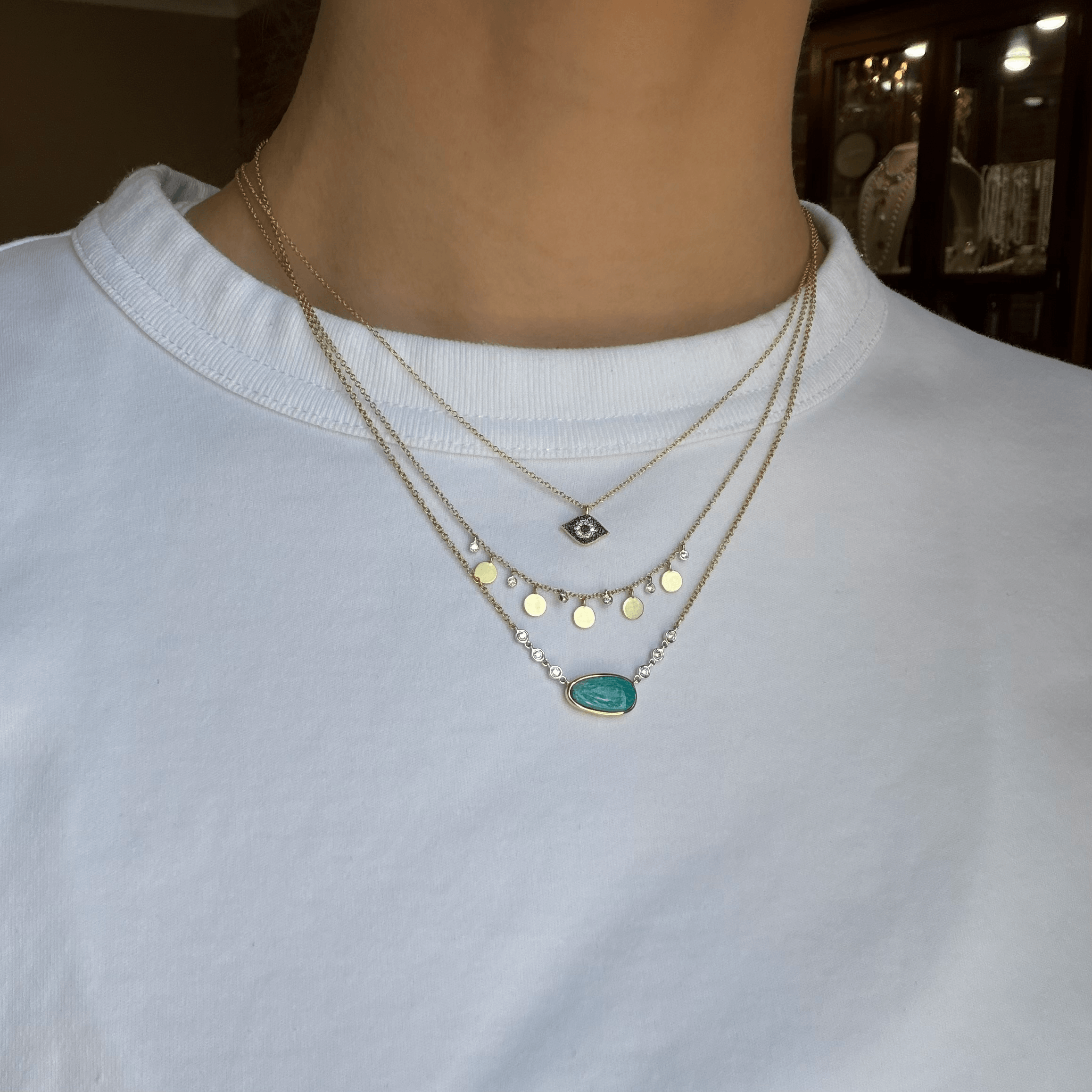 A close-up wearing a layered Meira T 14kt gold necklaces. The necklaces feature gold chain with an evil eye pendant, a second chain with gold disc charms, and a third chain with an oval opal pendant.
