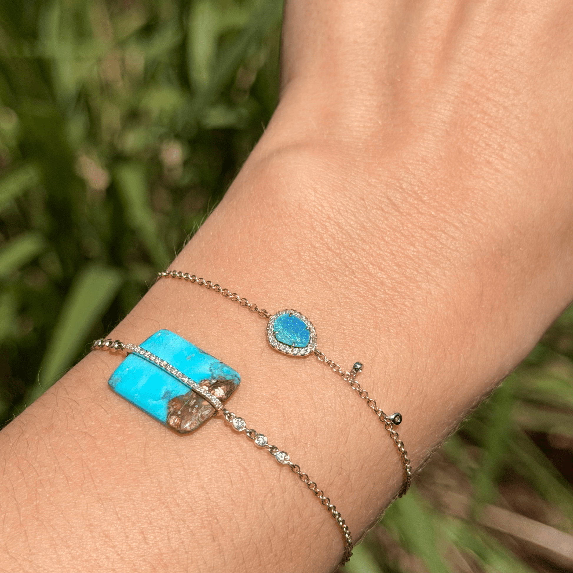 A close-up of a person's wrist wearing two Meira T 14k gold bracelets outdoors. One bracelet features a rectangular turquoise stone, and the other has a small oval opal stone surrounded by diamonds at Lindy's jewelry